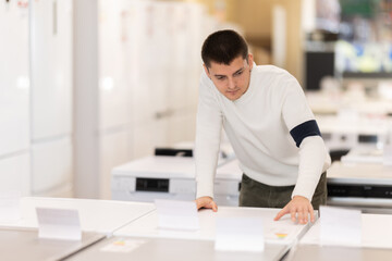 Guy buyer examines dishwashing machine or washing machine in detail before buying, checks completeness of household appliances and presence of defects. Customer chooses household appliances in store
