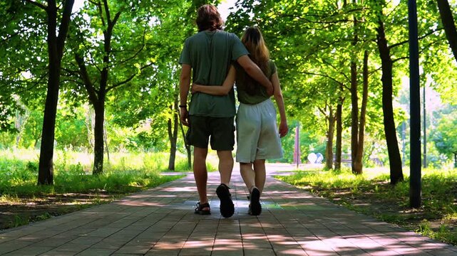 young loving couple holding hands walking towards camera along tree lined paved path in sunny summer park surrounded by lush green foliage dappled sunlight filtering through canopy creating romantic