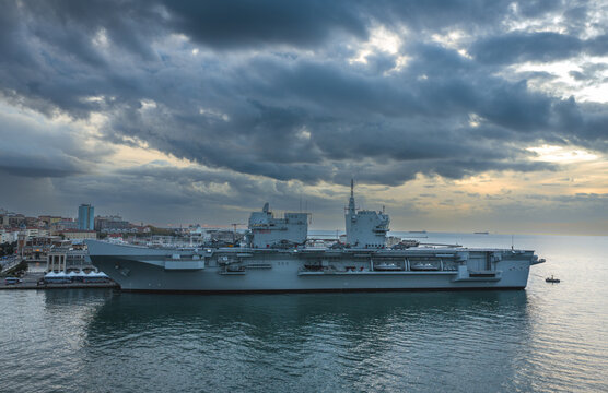 Aerial view of the imposing grey naval ship contrasts sharply with the ominous, gathering storm clouds over the still, reflective waters, Trieste, Italy.