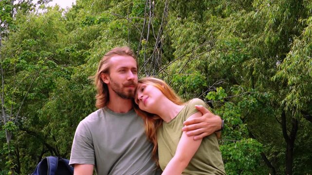 tender embrace of young loving couple outdoors in lush green summer forest man with long hair beard holding woman with reddish hair as she leans head on his chest both smiling gently eyes closed