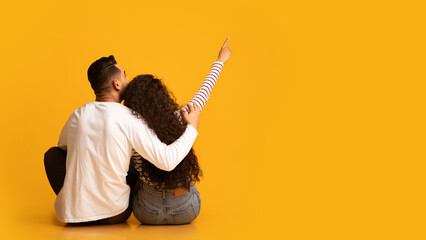 Rear view of young arab couple embracing and pointing up while sitting on floor over yellow studio...