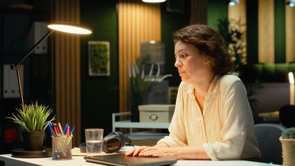 Overburdened woman prepares to leave office by turning off the lamp, doing overtime late at night due to intense workload and pressure. Uninterested discouraged worker. Camera B.