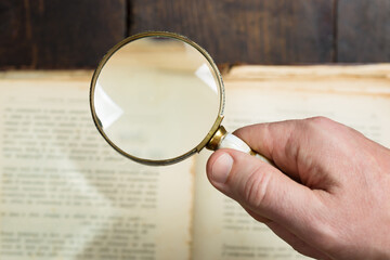 A hand holds a magnifying glass over printed text in an aged book.  Investigation, research of ancient documents.
