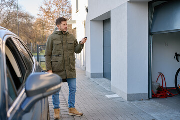 Man standing outside house near parked car using remote control to open garage door