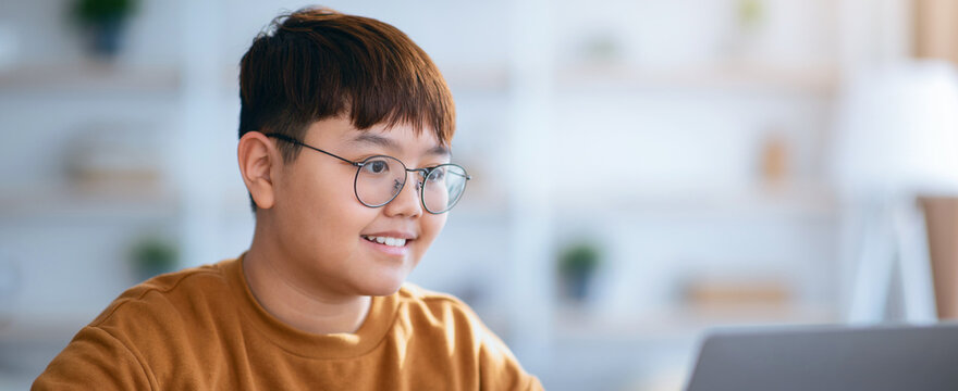 A boy is sitting at a table in his home, focused on his laptop screen. He is wearing glasses and has a light brown sweater. Books and plants are visible in the background. - Powered by Adobe