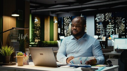 African american guy reviewing market data and building strategy for business goals, using spreadsheets on laptop for proofreading. Overtime after hours at multinational company.