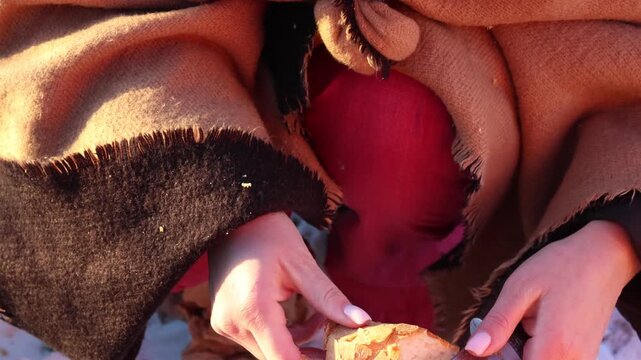 A close up shows hands slicing bread with a knife in the outdoors during winter, preparing food against a snowy backdrop, capturing the charm of rustic, wintry life.