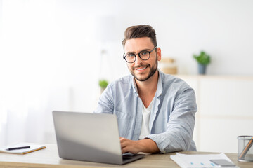 Fototapeta premium Portrait of freelancer man sitting at desk with laptop computer at home office, looking and smiling at camera, free space. Happy entrepreneur guy enjoying remote work and self-employment