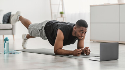 Online Workout. African American Fitness Man Exercising At Laptop Standing In Plank And Doing Leg Raises At Home. Black Millennial Male Training Near Computer In Living Room. Selective Focus