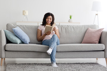 Fototapeta premium Study at home, education alone, planning day with notepad and smart hobby in spare time. Satisfied nerdy young african american lady read book on sofa, in minimalist living room interior, empty space