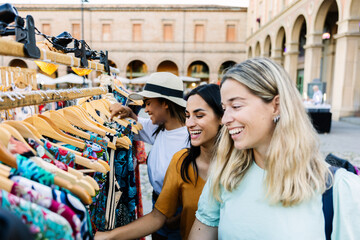 Multiracial young female friends choosing clothes outdoors. Cheerful women enjoying shopping time during a city trip. Lifestyle and travel concept.