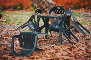 Stacked chairs and a table sit on the ground surrounded by fallen leaves. This outdoor area shows signs of a recent gathering or change in weather conditions
