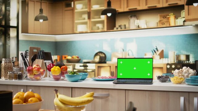 Green screen on laptop in modern home kitchen with cupboards and cookware on display. Countertop holds fresh organic produce and condiments, ideal background for cuisine or healthy eating.