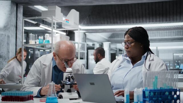 Pathologist examining biopsy slide while coworker documents diagnostic findings on laptop. Medical specialists working together to identify disease markers in clinical lab using notebook, camera A