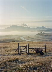 Rustic wooden fence and wheelbarrow filled with branches in a vast, frosty field, overlooking a winding river and misty mountains under a serene morning sky.