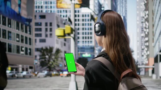 Vertical chroma key smartphone held by young adult with headset and backpack, gen z hipster girl walking the busy business district streets. Casual pedestrians of different ages. Camera A.