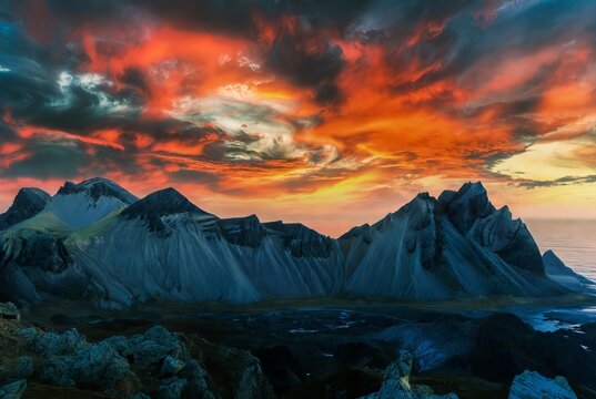 A breathtaking landscape featuring a jagged, dark mountain range under a chaotic and fiery sky - Powered by Adobe