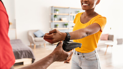 Smart Payment Concept. Closeup Of Courier Giving Terminal To Black Female Customer Paying With Smartwatch For Delivered Package Standing At Home. Selective Focus, Cropped Shot, Panorama