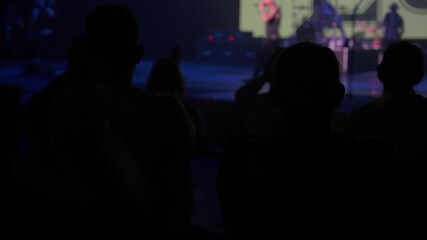 People stand in silhouette and raise hands while the stage and drums appear blurred in the background. Blue lights and smoke create a lively concert atmosphere inside the venue.