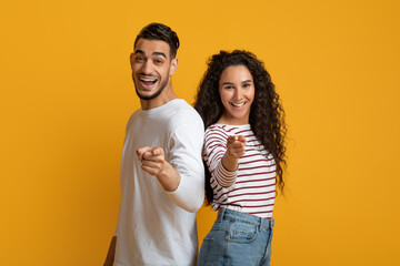 Obraz premium Joyful young arabic couple pointing at camera with fingers, cheerful middle eastern man and woman having fun while indicating somebody, posing together over yellow studio background, copy space