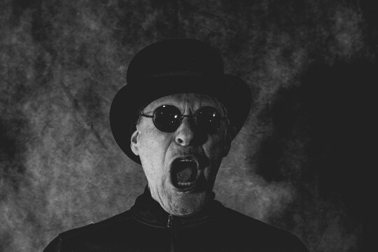 Black and white artistic portrait of a man in top hat and round glasses screaming intensely. Dramatic image ideal for concepts of madness, theater or psychological horror.