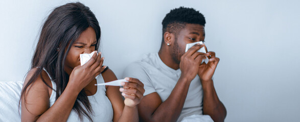 A couple sits on a bed while appearing unwell. They hold tissues and a thermometer, showing signs...