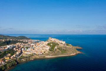 Fototapeta premium High angle view of Castelsardo's old town stretching along a dramatic peninsula, surrounded by deep blue Mediterranean waters and lush hills under a bright clear sky.
