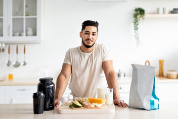 Portrait of handsome young Arab guy with healthy products and protein shake posing at kitchen....