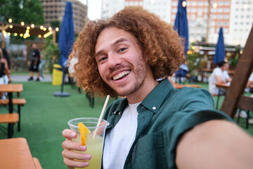 Young man with curly hair smiling and taking selfie while drinking a cocktail at open air bar