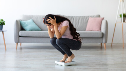 Young Indian woman sitting on scales, upset over weight gain at home, copy space. Emotional Eastern...