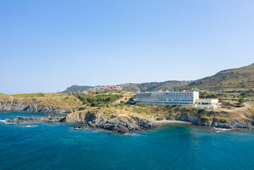 A contemporary hotel building stands on rugged coastal cliffs above turquoise Mediterranean waters near Banyuls-sur-Mer, with rolling hills and a distant village under a clear blue sky.