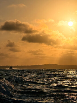 Golden-hour sunset over the Tapero&aacute; Channel in Bahia, Brazil, with a motorboat wake in the foreground, choppy water, and distant boats on the horizon.