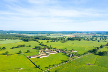 Wide aerial view of a traditional farmstead surrounded by green pastures and scattered trees near Cuncy les Varzy, with rolling hills and fields stretching to the horizon under a bright blue sky.