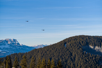 Two helicopters soar over a densely wooded mountain ridge with snowy peaks in the distance, under a clear blue sky in the French Alps. Bright winter sunlight highlights the rugged landscape and © Florent