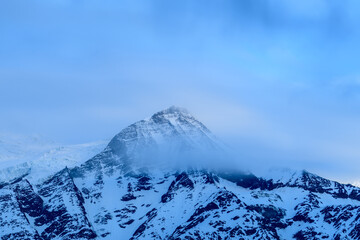 A snow-covered mountain peak emerges through drifting mist, illuminated by soft blue morning light in the Mont Blanc massif. Subtle textures and atmospheric clouds create a serene, tranquil mood.
