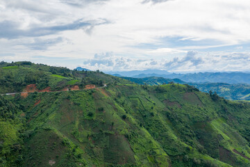 Naklejka premium A narrow mountain road curves along steep, lush green hillsides with distant blue ridges under a partly cloudy sky in the rural region between Phonsavan and Phou Khoun, Laos. The landscape features