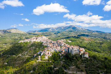 Obraz premium Aerial panorama of Rivello, Italy, showing a cluster of historic houses perched on a green hillside with dramatic mountains in the background. Bright daylight and scattered clouds create a vibrant