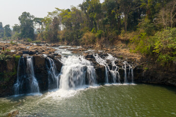Fototapeta premium Multi-tiered Tad Lo waterfall cascades over dark rocks into a wide green pool, bordered by dense forest and dry-season foliage in southern Laos. Warm sunlight highlights the flowing water, rugged