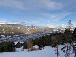 Sunny day view from a hillside in the Dolomites of a country house, its beautiful orange larch tree, the city of Brixen, Italy below with snow on all and the surrounding mountains in late November.