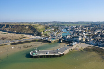 High-angle drone image of Port en Bessin showing the harbor entrance, surrounding piers, and clustered village buildings bordered by dramatic cliffs and open countryside under a clear sky.
