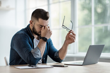 Tired freelancer works at home. Frustrated young man in shirt, with laptop at remote job, takes off...