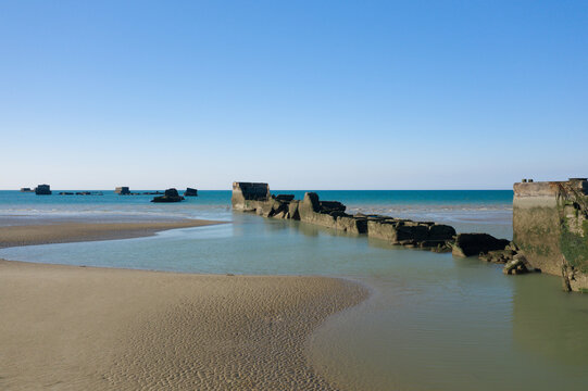 Concrete ruins of a historic artificial port stretch into calm turquoise waters at low tide, with smooth sand and a clear blue sky creating a tranquil coastal scene near Asnelles, Normandy.