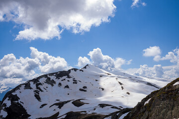 A sweeping view of a snow-covered mountain ridge on Aiguillette des Houches, highlighted by textured rock outcrops and billowing cumulus clouds under a vibrant blue sky.