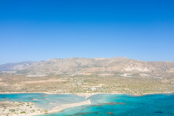 Obraz premium Wide aerial image of Elafonisi beach in Crete, Greece, featuring turquoise waters, pale sandy stretches, and rugged, sun-bleached hills beneath a clear blue sky. The atmosphere is bright, open, and