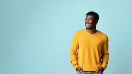 Happy millennial black man in casual outfit holding hands in pockets, looking at copy space for text or advertisement and smiling, recommending nice offer or discounts, studio shot on blue, panorama © Prostock-studio