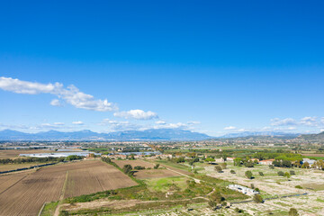Obraz premium Expansive rural landscape with cultivated fields, archaeological remains, and scattered buildings stretching toward distant mountain ranges under a vivid blue sky with scattered clouds. The scene