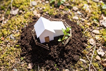 Small white paper house model on soil mound with green plant sprout and wooden fence outdoors