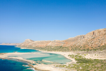 Shallow turquoise lagoons meet rugged rocky cliffs at Balos Beach in Crete, Greece, under a clear blue sky. The scene is sunlit, natural, and expansive, with contrasting textures and vibrant colors.