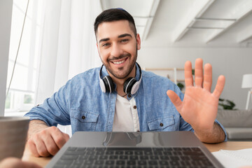 Headshot portrait of happy young 30s man sitting at desk at home office, looking at camera, waving hello. Excited teacher lecturer recording educational video, guy greeting students at online workshop
