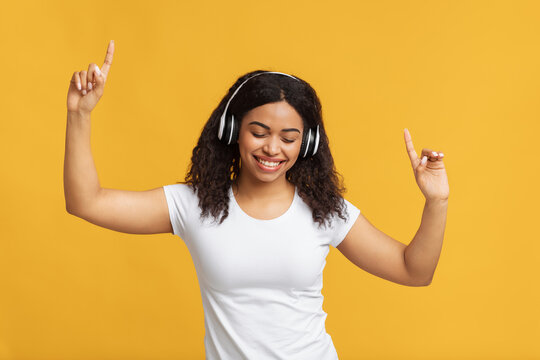 Carefree african american lady in headphones dacing to her favorite playlist with closed eyes over yellow studio background. Black woman moving to beautiful song. Pastimes and hobbies concept